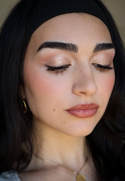 Primer plano de un rostro con piel de tono marrón claro, cabello oscuro, cejas definidas, maquillaje de ojos brillante y labios nude brillantes. Pendientes de aro dorados y collar visibles.