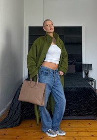 Femme portant un manteau vert olive, un crop top blanc, un jean ample et des chaussures à enfiler à carreaux, tenant un sac à main beige, debout dans une chambre.