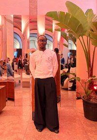 Egy személy áll egy tükrözött térben, fehér hosszú ujjú inget visel, melyen apró minta látható, valamint széles fekete nadrágot, előtérben egy növénnyel.