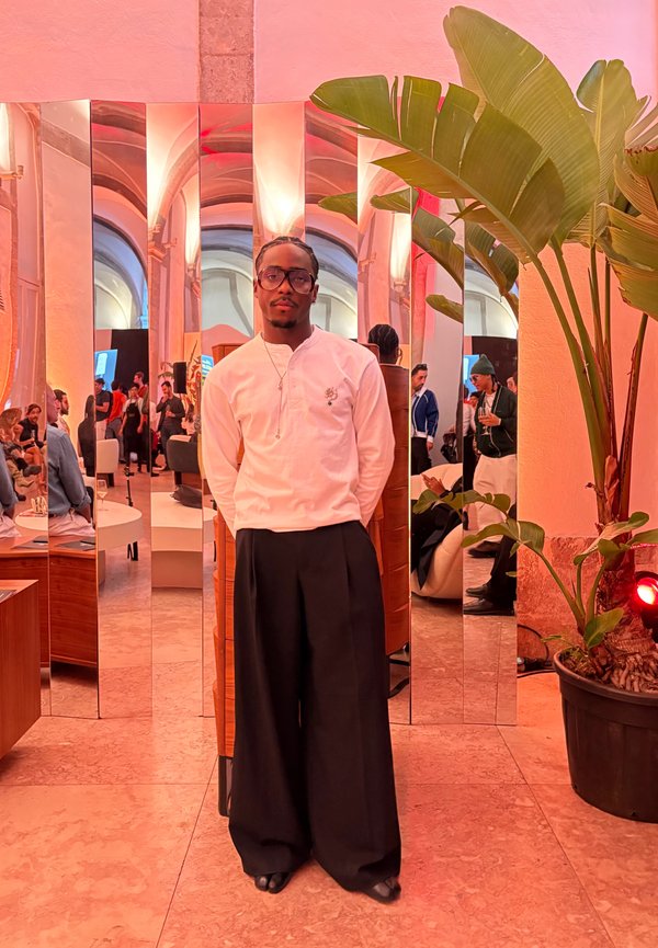 Egy személy áll egy tükrözött térben, fehér hosszú ujjú inget visel, melyen apró minta látható, valamint széles fekete nadrágot, előtérben egy növénnyel.