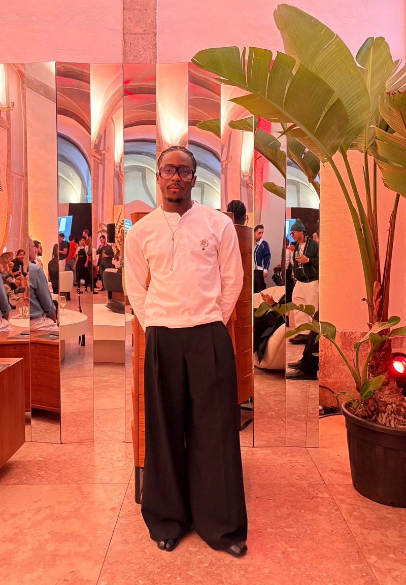 Une personne se tient dans un espace aux murs miroirs, portant une chemise blanche à manches longues avec un petit motif et un pantalon large noir, une plante est visible au premier plan.