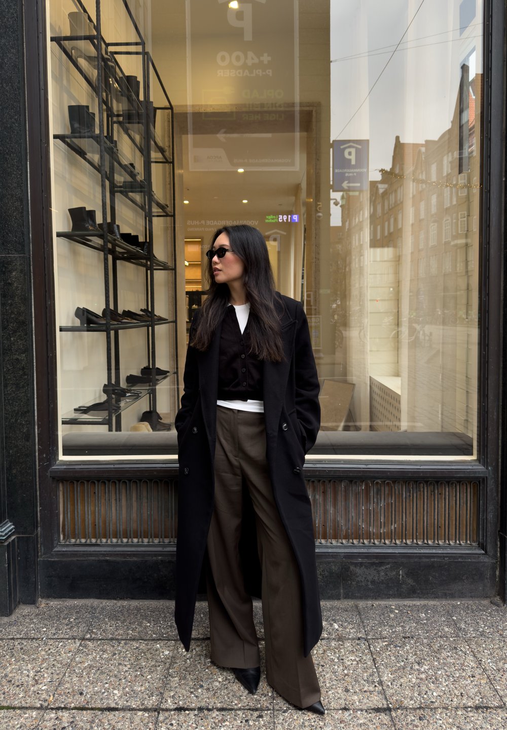 Manteau noir croisé à double boutonnage porté sur un gilet noir ajusté, associé à un pantalon large marron et des chaussures noires à bouts pointus, debout devant une boutique.