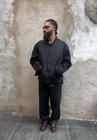 Schwarze gepolsterte Jacke mit Knopfverschluss, über einem schwarzen Shirt getragen; kombiniert mit dunklen, weit geschnittenen Hosen und burgunderfarbenen Schuhen. Glatte Textur.