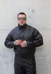 Svart skinnjakke med høy krage og elastisk midjebånd, kombinert med svarte skinnbukser. Modellen har på seg mørke solbriller og har tatoveringer.