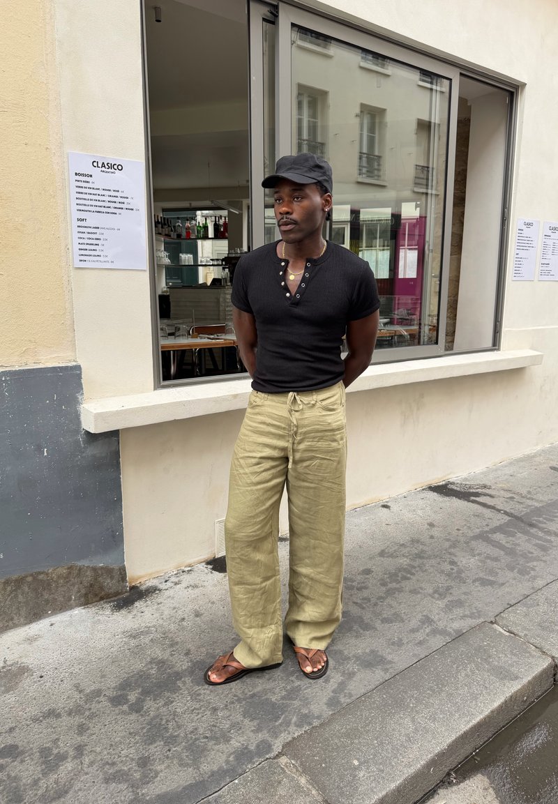 Homme portant une chemise noire, un pantalon beige et des sandales se tient les mains dans le dos devant la fenêtre d'un café avec un menu sur le mur.
