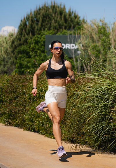 Atleta con un sujetador deportivo negro y pantalones cortos blancos, corriendo por un camino, usando gafas de sol tintadas en azul y zapatillas deportivas moradas.