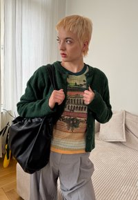 Cardigã verde e fofinho sobre um suéter canelado com padrões em tons terrosos. Uma bolsa preta e macia e calças largas cinzas completam o look.