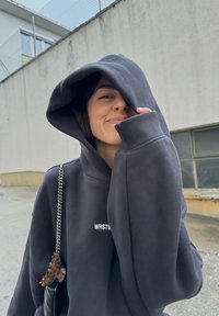 Μαύρη hoodie με μεγάλο, μαλακό καπέλο και κεντημένο λογότυπο "WRSTB". Ένας τσάντα με αλυσίδα και τονισμένα στοιχεία από χελώνα είναι ορατή.