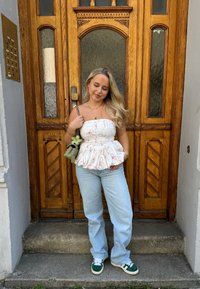 Weißes Peplum-Top mit pinkem Wirbelmuster, hellblaue Jeans, grüne und weiße Sneaker, steht vor einer Holztür mit Glasfenstern.