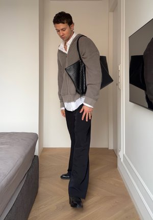 Cardigan gris de punto sobre una camisa blanca, pantalones negros de pierna ancha y zapatos negros. Sosteniendo un bolso tote negro con textura, de pie en una habitación.