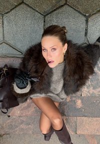 Brun pälsjacka över en grå tröja, svarta läderhandskar, håller solglasögon. Mörk handväska bredvid. På en strukturerad stenyta.