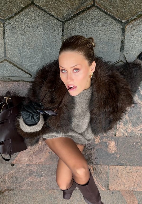 Brun pelsfrakke over en grå sweater, sorte læderhandsker, holder solbriller. Mørk håndtaske ved siden af. På en tekstureret stenovertflade.