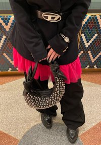 Zwarte handtas met een gebogen vorm, versierd met zilveren studs. Beschikt over een geknoopte handgreep, samen met een roze gelaagde rok en zwarte broeken.