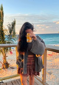 Γυναίκα με μακριά μαλλιά που καλύπτουν το πρόσωπό της κρατάει ένα κίτρινο κουτί ψηλά, στέκεται σε ξύλινη εξέδρα στην παραλία με ωκεανό και φοίνικες στο φόντο.