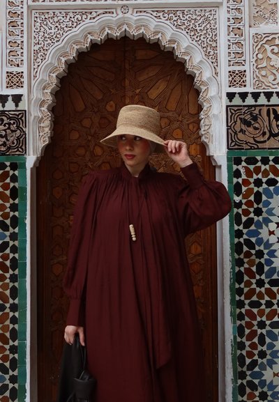 Mujer con vestido granate y sombrero de paja ajustando el ala, de pie frente a una puerta de madera ricamente tallada con un marco detallado de azulejos de mosaico.