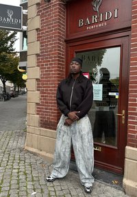 Fekete zipzáras bombázó dzseki, világosszürke, oversize farmernadrág, fakó mintával, és fekete-fehér sneaker cipők, egy bolt előtt állva.