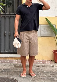 Cămașă polo neagră, pantaloni scurți bej, sandale; ținând o pălărie de o culoare deschisă. Fundalul prezintă o ușă și o plantă verde într-un ghiveci.