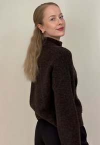 Jeune femme aux longs cheveux clairs attachés en arrière, portant des boucles d'oreilles dorées, un pull foncé et duveteux, ainsi qu'un pantalon noir, légèrement tournée et souriante.