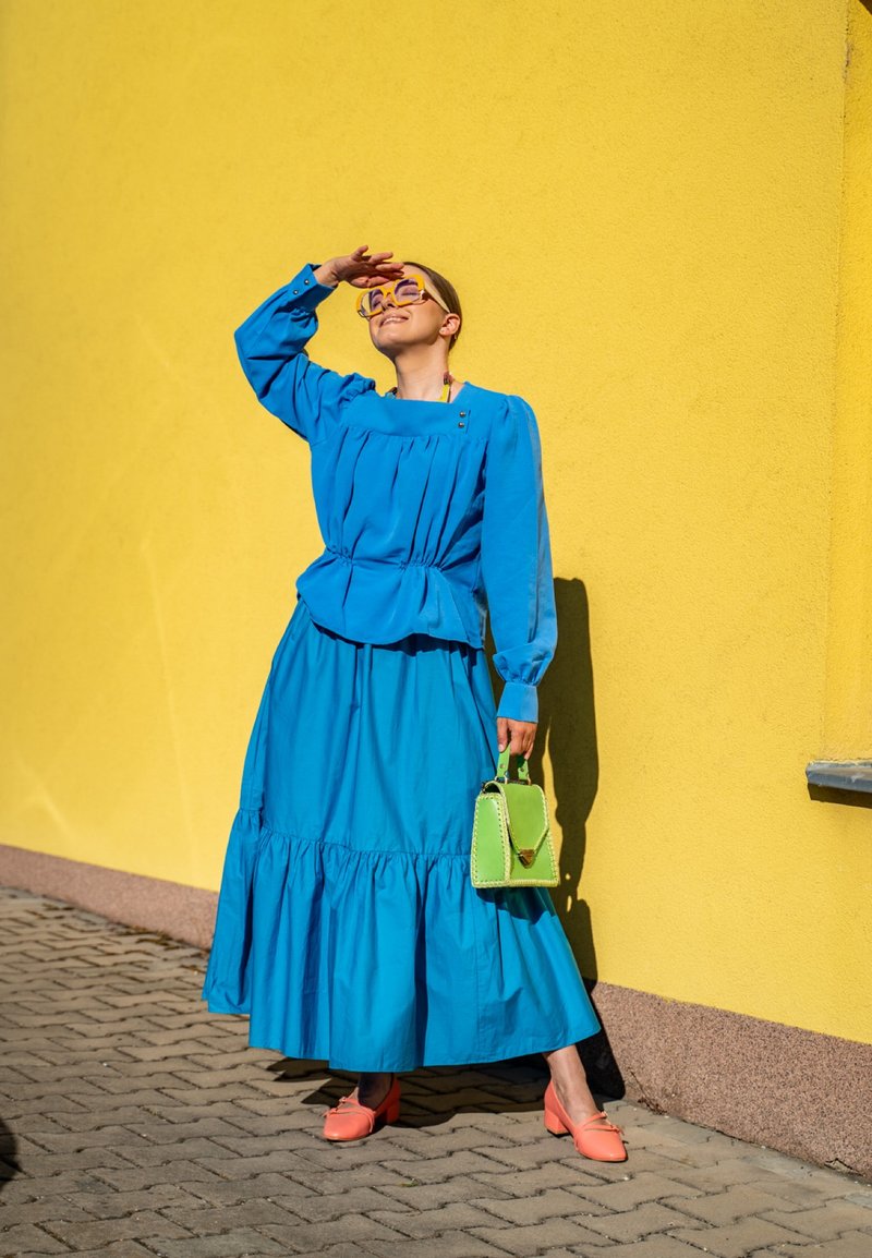 Femme en robe bleu vif et chaussures orange debout contre un mur jaune, se protège les yeux avec la main et tient un petit sac à main vert.