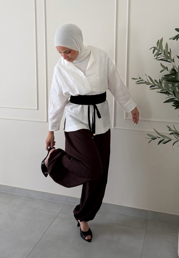 Femme portant un hijab blanc et un chemisier avec une ceinture noire ajuste une chaussure noire à talon haut tout en se tenant sur une jambe dans une pièce minimaliste.