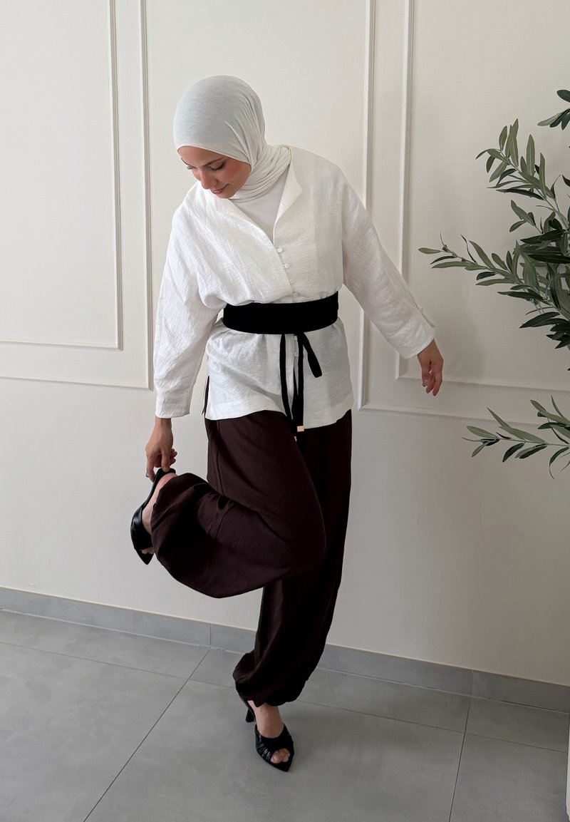 Femme portant un hijab blanc et un chemisier avec une ceinture noire ajuste une chaussure noire à talon haut tout en se tenant sur une jambe dans une pièce minimaliste.