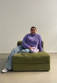 Grønn kordfløyelssofa med buet rygg og bred sete. En person i en lilla, strukturert genser sitter avslappet og holder en liten lavendelpose.