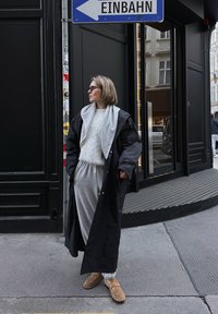 Fekete, oversized kabát szürke kapucnis pulóverrel alatta, szürke melegítőnadrág és bézs papucs. Városi háttér modern építészetel és utcai táblákkal.