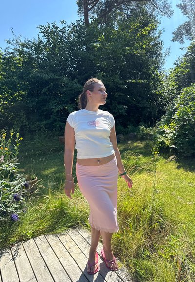 Camiseta corta blanca con un gráfico rosa, falda midi rosa y sandalias rosas. Entorno exterior soleado y cálido con vegetación.