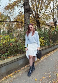 Lichtblauw geribde trui, witte asymmetrische rok met een textuurontwerp, zwarte platform veterschoenen en een zwarte handtas. Bladeren verspreid op de grond.