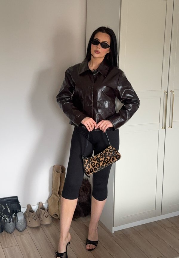 Femme portant une veste en cuir sombre, des leggings noirs courts, des sandales à talons noires, tenant un sac à main imprimé léopard, debout dans une pièce avec des chaussures et une armoire.