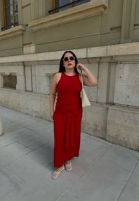 Robe rouge, côtelée, sans manches avec un d étail de taille nouée, accompagnée de chaussures plates transparentes et d'un sac à main crème, le tout devant un mur en pierre.