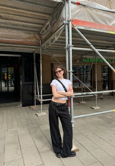 Pantalones de pierna ancha negros, camiseta cortada blanca y zapatos chunky negros. De pie contra andamios, con una bolsa de bandolera y gafas de sol.