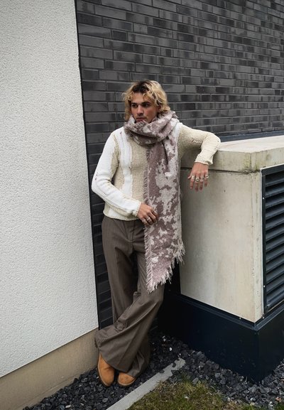 Suéter grueso de crema con tejido texturizado, pantalones ajustados de raya marrón y una bufanda rosa con estampado. Los accesorios incluyen múltiples anillos.