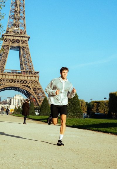 Corredor con una chaqueta ligera y translúcida con cremallera y pantalones cortos negros, troteando por un sendero con la Torre Eiffel de fondo, bajo cielos azules despejados.