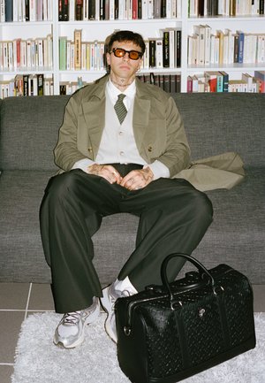 Schwarze Leder-Duffel-Tasche mit strukturiertem Muster und Hardware-Details, kombiniert mit locker sitzenden dunklen Hosen und grauen Sneakers, auf einem grauen Sofa.