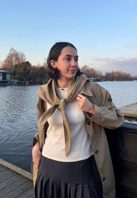 Μπεζ παλτό με υπερμεγέθη γιακά, κρεμαστή κασκόλ, λευκή υφασμάτινη μπλούζα και ναυτικό πλισέ φούστα. Νερό στο φόντο.