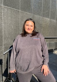 Sudadera grande de color gris oscuro con puños acanalados y cuello redondo, con los textos "Studio Uth" y "TRAVEL BASICS". Accesorio: bolso negro con tachuelas.