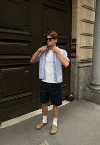 Młody mężczyzna w okularach przeciwsłonecznych, białej koszulce, paskowanej koszuli z krótkim rękawem, ciemnych szortach, białych skarpetkach i beżowych klapkach stoi przy dużych drewnianych drzwiach.