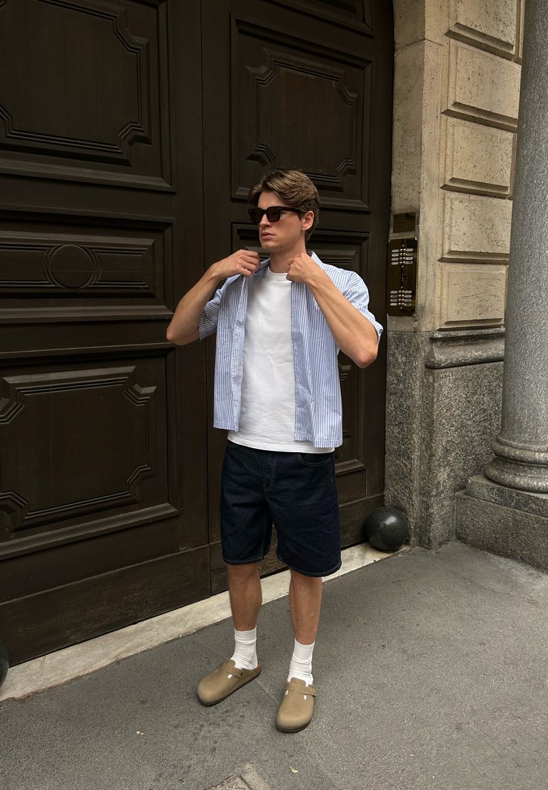 Giovane uomo con occhiali da sole, t-shirt bianca, camicia a righe a maniche corte, pantaloni corti scuri, calze bianche e zoccoli beige in piedi accanto a una grande porta di legno.