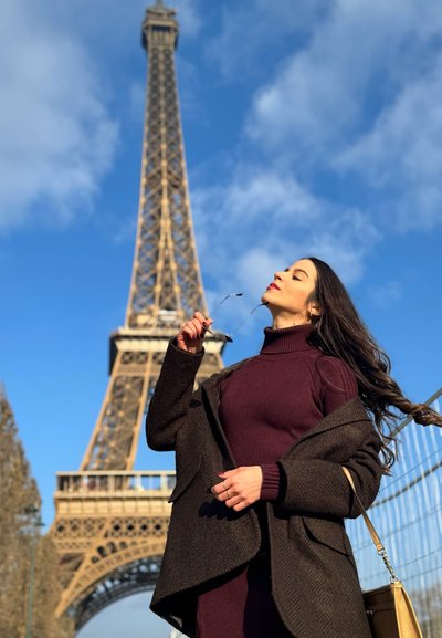 Una mujer con un abrigo marrón oscuro y un suéter burdeos sostiene unas gafas de sol, de pie frente a la Torre Eiffel contra un cielo azul.