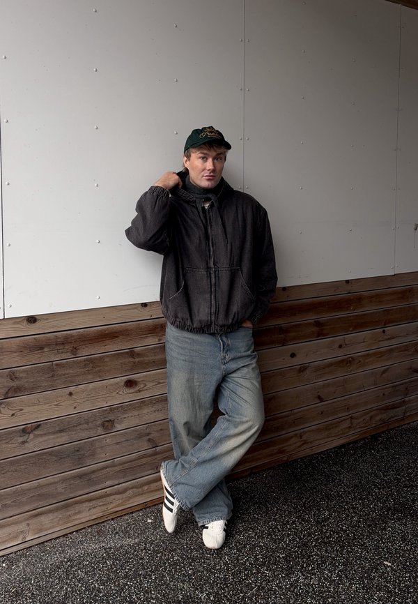 Jeune homme portant une veste noire, une casquette verte, un jean bleu et des baskets blanches, appuyé contre un mur en panneau de bois et blanc.