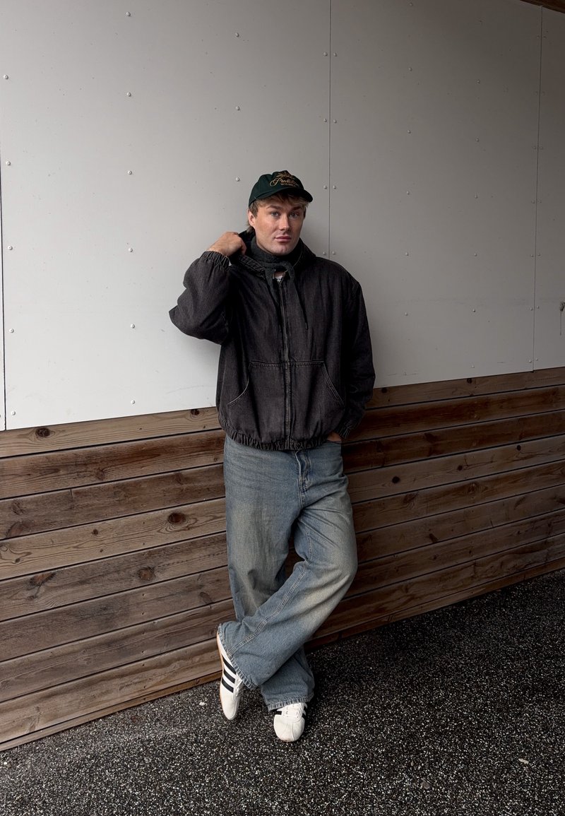 Jeune homme portant une veste noire, une casquette verte, un jean bleu et des baskets blanches, appuyé contre un mur en bois et blanc.
