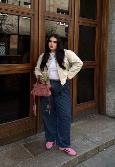 Chaqueta bomber beige, top recortado blanco, jeans de pierna ancha azules, zapatillas rosas con rayas negras y un bolso rosa, sosteniendo una bebida.