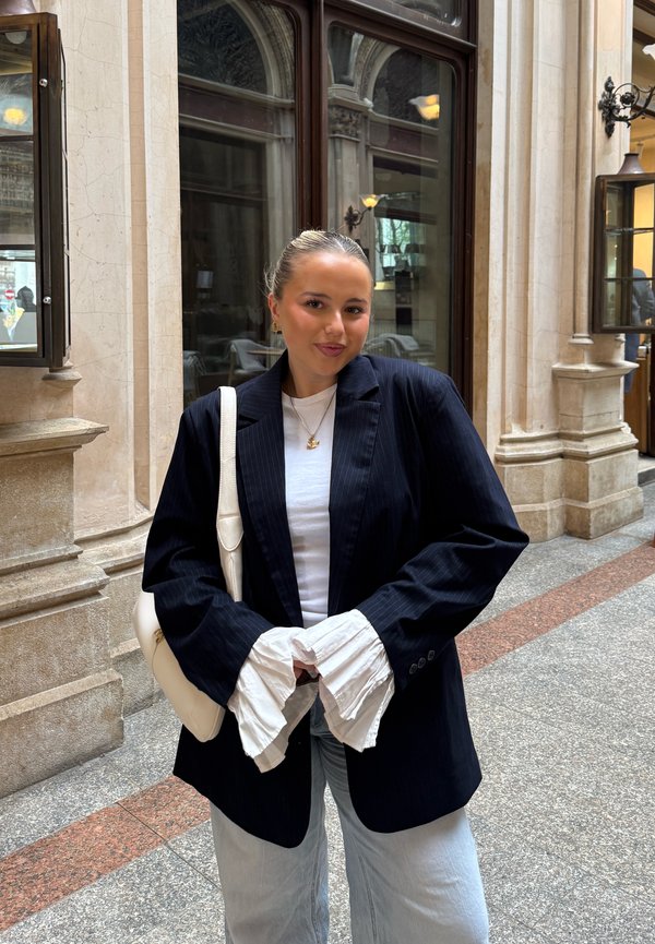 Jeune femme aux cheveux lissés en arrière, portant un blazer noir sur une chemise blanche et un jean clair, debout dans un couloir en pierre avec des fenêtres en arc.