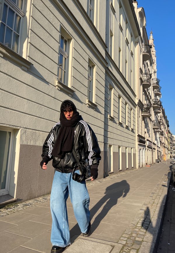 Yli suuri musta nahk taku, jossa on valkoiset raidat, musta huivi, vaaleansiniset leveät housut ja mustia asusteita, kävelee jalkakäytävällä.