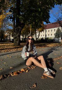 Bluză sport cu design grafic, pantaloni scurți negri, șosete albe, adidași maro. Ținând o cană neagră de cafea, așezat printre frunzele de toamnă.