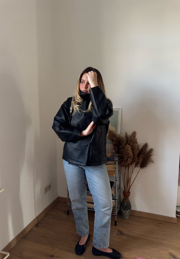 Fiatal nő fekete dzsekiben és világoskék farmerben áll egy beltéri polc mellett, amelyen könyvek és egy váza szárított növényekkel található.
