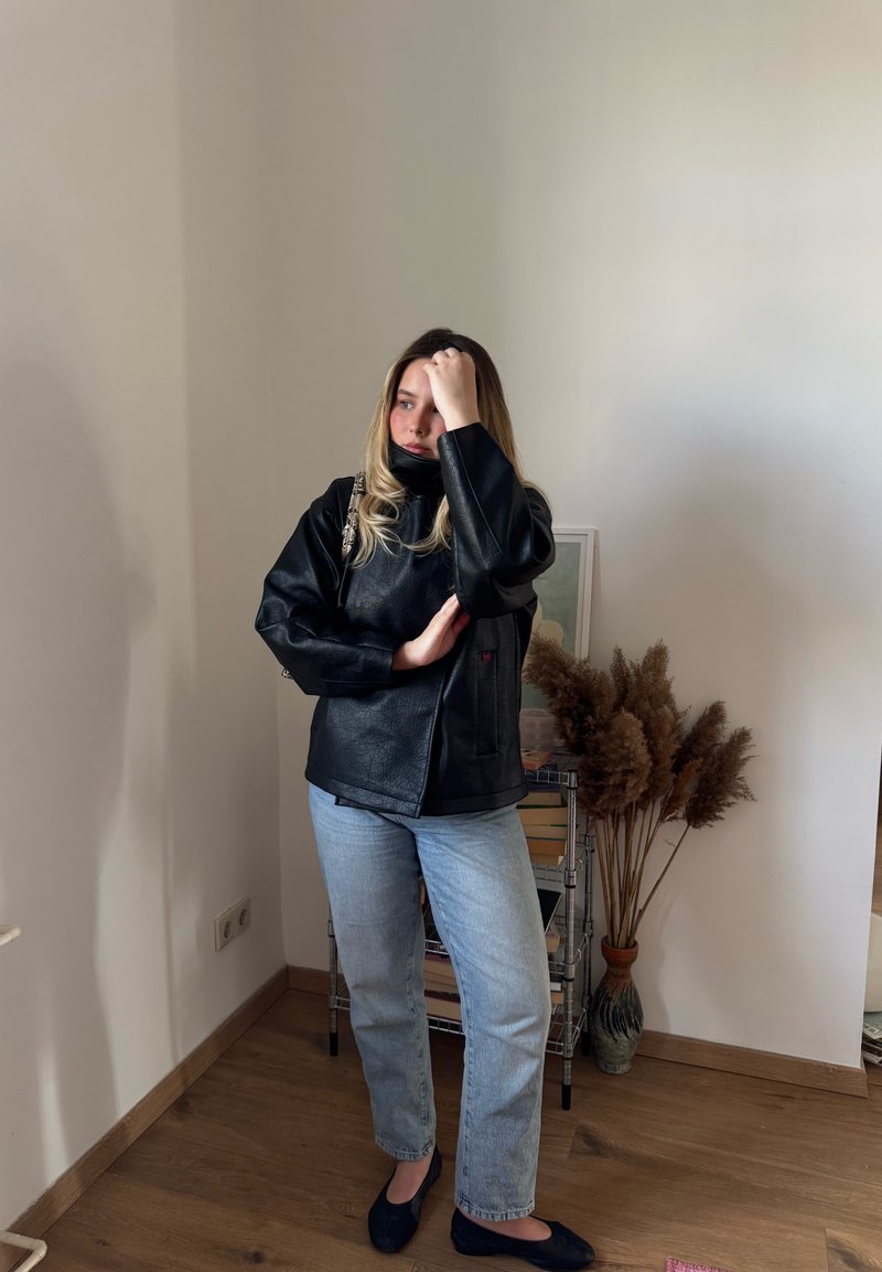 Jonge vrouw in een zwart jasje en lichtblauwe jeans staat binnen bij een plank met boeken en een vaas met gedroogde planten.