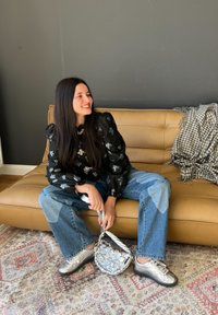 Fekete virágmintás blúz puffos ujjakkal, világoskék foltos farmernadrág, ezüst sneaker cipők, és fényes, virágmintás vödrös táska.