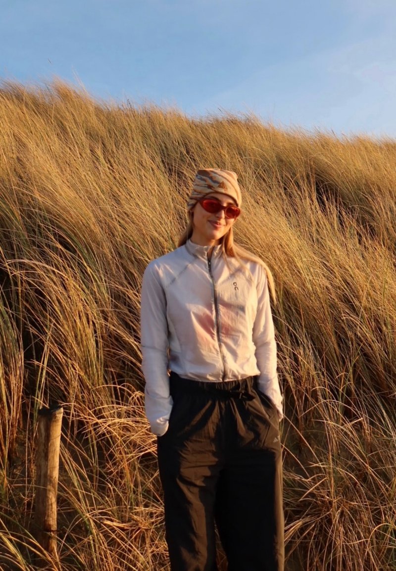 Femme portant des lunettes de soleil, une veste légère et un foulard à motifs, debout dans une haute herbe dorée sous un ciel bleu clair.